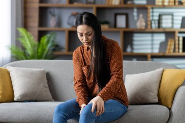Asyalı kadın kanepede oturmuş dizine bakıyor ve rahatsızlığını dile getiriyor. Sakin bir ev ortamında günlük giysiler içinde. Acı, rahatlama, ev, sağlık, sağlık, yaşam tarzı kavramı.