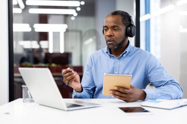 Video konferansında, kulaklık takan, dizüstü bilgisayarla konuşurken not defteri tutan olgun Afro-Amerikan iş adamı. Modern ofiste sanal bir buluşmayla meşgul. Uzaktan çalışmayı, teknolojiyi temsil eder.