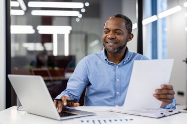 Modern ofis ortamında dizüstü bilgisayarla iş belgelerini inceleyen olgun bir Afro-Amerikan işadamı. Kendine güvenen, odaklanmış, mavi gömlekli, gülümseyen bir profesyonel..