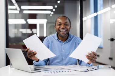 Olgun afro-amerikan iş adamı güvenle gülümsüyor, modern ofis ortamında kağıtları tutuyor. İş ortamında başarı, pozitiflik ve profesyonelliği temsil eder.