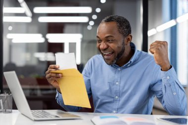 Olgun Afrikalı Amerikalı iş adamı başarıyı kutluyor. Dizüstü bilgisayarın yanında mutluluk ve başarıyı ifade eden bir zarf, neşeyi simgeliyor.
