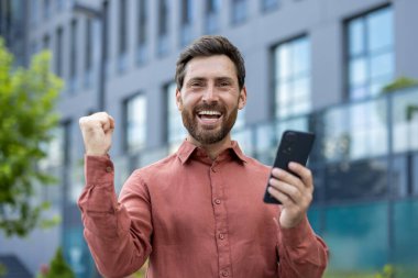 Olgun bir adam modern binanın yakınında akıllı telefon tutmaktan zevk alıyor. Başarıyı kutluyoruz, olumlu haber. Sıradan tarz, iş zaferi, şehir çevresi, neşeli ve heyecanlı.