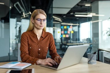 Ofiste dizüstü bilgisayarla çalışan ciddi bir iş kadını. Olgun profesyoneller önemli görevlere odaklanır. İş kıyafetleri ve örgütlü çalışma alanı modern çevrede özveri ve uzmanlığı vurguluyor.