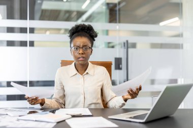 Genç Afrikalı Amerikalı iş kadını masasına şaşkın şaşkın bakıyor. Bilgisayarlı ofis ortamı, belgelere göre iş ortamında stres ve karar verme süreci.