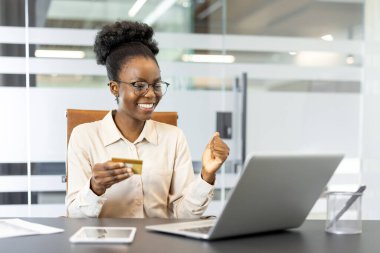 Gülümseyen Afro-Amerikan iş kadını ofis masasında bilgisayarını online alışveriş için kullanıyor. Kredi kartının olması e-ticaret, finans ve iş ortamında başarılı olduğunu gösteriyor. Telefon masada görünür..