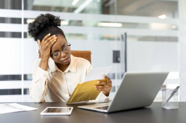 Afro-Amerikalı iş kadını ofiste belgeleri incelerken stresli görünüyor. Dizüstü bilgisayarının yanında akıllı bir telefon kullanıyor. Baskı ve karar verme duygularını iletiyor..