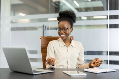 Genç Afrikalı Amerikalı iş kadını ofisinin masasında neşeli bir şekilde meditasyon yapıyor. Sanal bir buluşmada dizüstü bilgisayar ve tablet kullanır, profesyonelliği somutlaştırır ve odaklanır..