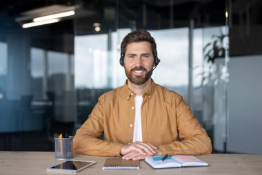 Kulaklık takan olgun bir işadamı ofis masasında rahatça oturur. Not defteri ve tablet gibi dijital teknolojiyle ilgilenir, profesyonelliği ve verimliliği tasvir eder.