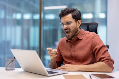İşadamı ofisteki dizüstü bilgisayarı işaret ederken hayal kırıklığı gösteriyor. İş sırasında öfke ya da korku gibi duyguları ifade eder. Modern ortamda kırtasiyeyle birlikte masada oturuyordu..