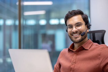 Kulaklık takan İspanyol işadamı ofis ortamında dizüstü bilgisayarla çalışıyor. Kendine güvenen gülümseme, kahverengi gömlek, gözlük, profesyonellik. Parlak, modern arka plan. İş ve iletişim.