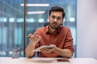 Modern ofis ortamında elinde bir defter tutarken kırmızı gömlekli bir iş adamı el kol hareketi yapar. Odaklanmış ifade iletişim, planlama ya da karar vermeyi önerir. Akıllı telefon masada görünür..