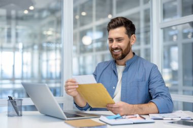 Modern ofiste, masadaki bilgisayarla mutlu bir şekilde zarfı açan adam olumlu duygu, iletişim ve iş başarısını gösteriyor. Şık dekorlu parlak ayar.