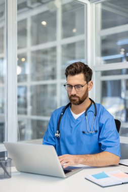 Mavi önlük giyen ve gözlüklü bir tıp uzmanı modern ofiste dizüstü bilgisayarla çalışıyor. Sağlık teknolojisini, teletıp ve sağlık hizmetlerini temsil ediyor. Ofis ortamı