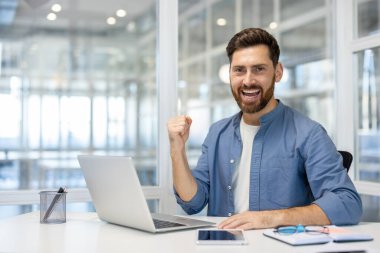 Modern ofiste başarıyı kutlayan gülümseyen adam, dizüstü bilgisayar ve telefonla masada oturuyor. Neşeli ifade başarıyı ve mutluluğu gösterir..