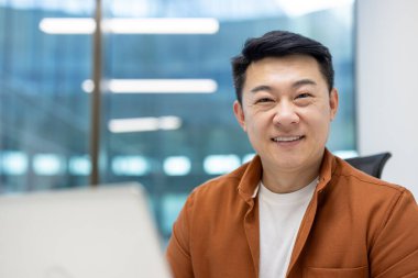 Asyalı iş adamı modern ofis ortamında dizüstü bilgisayarla güvenle gülümsüyor. Geniş pencereli parlak profesyonel ortam, rahat çalışma alanı ve verimliliği vurgulayan günlük kıyafet.