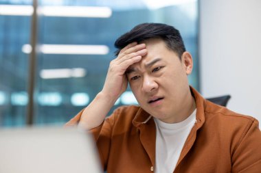 Asyalı iş adamı dizüstü bilgisayarla uğraşırken stresli hissediyor. Teknoloji ve zorluklarla dolu ofis ortamı. Baskı ve sorun çözme zihniyetini gösteren iş ortamı.