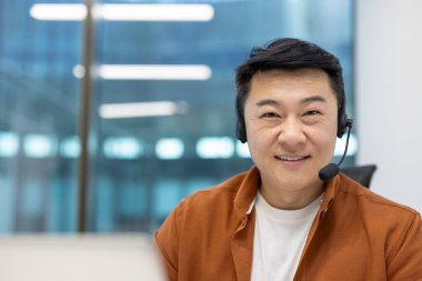 Modern ofiste gülümseyen kulaklık takan Asyalı bir iş adamı. Video ya da telekonferansa profesyonel katılımın göstergesi. Güven, dijital iletişim ve çalışma ortamını betimler.