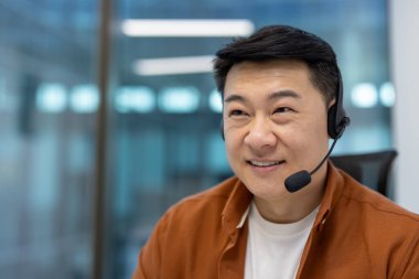 Asyalı iş adamı modern ofiste masasına gülümsüyor, internet iletişimi için kulaklık takıyor. Parlak, odaklı ifade pozitifliği ve dijital iş toplantısına etkin katılımı öneriyor.