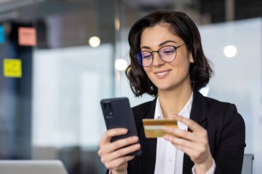 Genç iş kadını akıllı telefon tutuyor, modern ofiste kredi kartıyla online alışveriş yapıyor. Dijital işlem, finans teknolojisi, iş yeniliği ve mobil güvenliği vurguluyor.