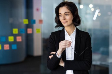 Ofisteki kendine güveni tam bir iş kadını, elinde gözlük tutuyor, profesyonel kıyafetler giyiyor. Liderlik kavramı, iş ortamında tefekkür. Canlı yapışkan notlar yaratıcı dokunuşlar ekler.