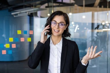 Çağdaş ofiste genç bir iş kadını telefonda konuşuyor. Profesyonel iletişim ve çoklu görev becerileri apaçık ortada. İş ortamında güven, iş görevlerinde uzmanlık.