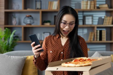 Evde telefonu tutan Asyalı kadın pizza siparişini heyecanla alıyor. Pizza ortaya çıktığında neşe, sürpriz ve hoşgörü anı yakalar, eğlence ve teknoloji bağlantısını vurgular.