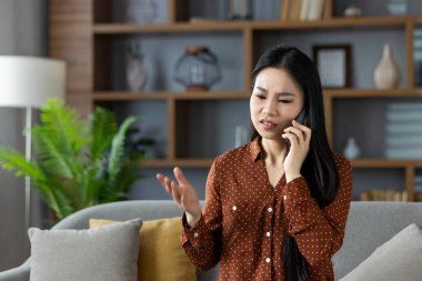 Telefondaki Asyalı kadın evde önemli bir konuyu tartışıyor. Rahat bir oturma odasında gri koltukta otururken kahverengi benekli tişört giymek..