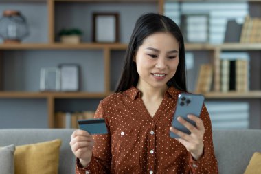 Asyalı bir kadın elinde kredi kartıyla online alışveriş için akıllı telefon kullanıyor. Modern yaşam tarzı, kolaylık ve teknoloji kullanımını yansıtan dijital işlemlerle iç içe.
