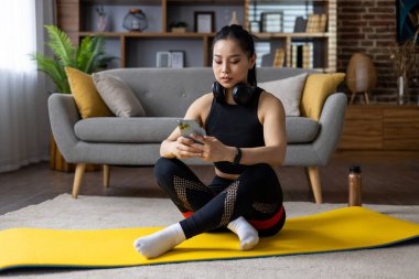 Yoga minderinde egzersiz yaparken akıllı telefon ve kulaklık kullanan Asyalı kadın. Çağdaş yaşam tarzını ve farkındalık uygulamalarını yansıtan fitness aktiviteleriyle meşgul.