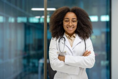Beyaz laboratuvar önlüğü giyen, hastanede kolları bağlı duran kendinden emin Afrikalı Amerikalı kadın doktor. Sağlık hizmetlerinde güvence ve uzmanlık sergileyen tıbbi profesyoneller.