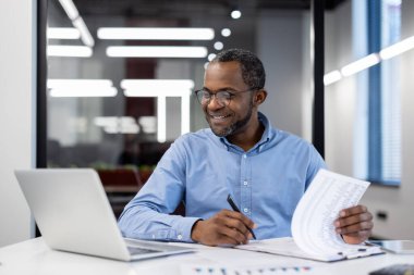 Olgun bir Afro-Amerikan işadamı modern ofisteki raporları analiz ediyor. Dizüstü bilgisayarda çalışan profesyonel bir adam, finansal belgeleri dikkatle inceleyen, kendine güvenen ve yüzünde bir gülümseme olan biri..