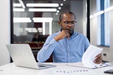 Olgun Afrikalı Amerikalı işadamı, modern ofiste mali belgeleri inceliyor. Odaklanmış ifade, eleştirel düşünme ve problem çözmeyi gösterir. Laptop ve evrak işleri profesyonel ortam.