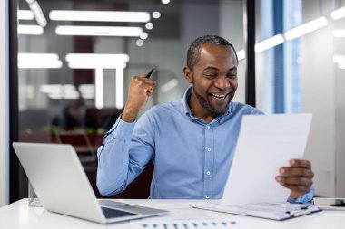 Olgun Afro-Amerikan iş adamı ofis masasında oturmuş başarıyı kutluyor. Belgeler, kalemler ve dizüstü bilgisayarlarla gülerken. Profesyonel başarı ve mutluluğu simgeliyor..