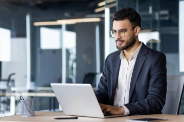 Modern ofiste dizüstü bilgisayar kullanan bir iş adamı, kurumsal göreve odaklanmış. Profesyonel kıyafet ve ciddi ifade biçimi, iş projesine olan bağlılığını ve konsantrasyonunu ifade eder. Gözlükler dikkat çekiyor.