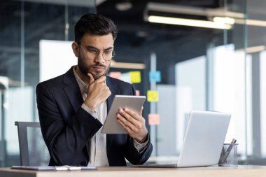 Modern ofisteki profesyonel işadamı, düşünceli bir ifade sergilemek için tablet ve dizüstü bilgisayar kullanıyor. Teknoloji, analiz, iş anlayışı. Resmi takım elbiseli, işyerinde görevli..