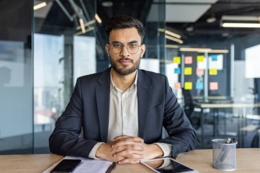 Modern ofisteki masasında oturan kendine güvenen iş adamı. Etrafı kağıt, kalem ve akıllı telefonla çevrili. Cam duvarlar ve renkli yapışkan notlar profesyonel ayarları güçlendirir.