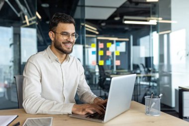 Modern ofis ortamında dizüstü bilgisayar kullanan gülümseyen profesyonel bir adam. Gözlük ve açık renk gömlek takmak, üretkenlik, teknoloji ve iş ortamı sergilemek. Pozitif ve odaklı işyeri.