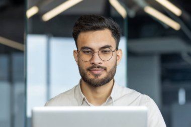 Modern ofis ortamında dizüstü bilgisayarda çalışan gözlüklü genç profesyonel bir adam. Teknoloji, iş ve üretkenlik kavramları önererek, göreve odaklanmış ve meşgul görünüyor.