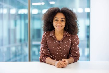 Modern ofisteki kendinden emin Afrikalı Amerikalı genç iş kadınının portresi. Şık bir benekli bluz giyiyor, iş ortamında profesyonellik, özgüven ve hırs gösteriyor. Doğal ışık.