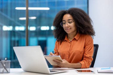 Modern ofisteki genç Afrikalı Amerikalı iş kadını dizüstü bilgisayar ve telefonla not alıyor. Gözlüklü ve turuncu gömlekli, parlak, profesyonel çalışma alanıyla çevrili..