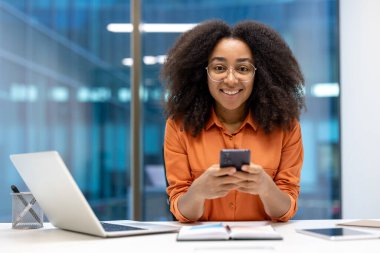 Ofisteki kendine güvenen Afrikalı Amerikalı iş kadını akıllı telefonunu masada kullanırken gülümsüyor. Modern işyeri teknoloji ve verimliliği ön plana çıkarıyor, telefon, dizüstü bilgisayar ve verimliliği ön plana çıkarıyor.