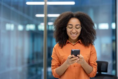 Modern ofiste akıllı telefon kullanan, iş arasında ekranda gülümseyen, gözlüklü, kendine güvenen genç bir iş kadını. Teknolojiden ve profesyonel yaşam tarzından anlıyor..