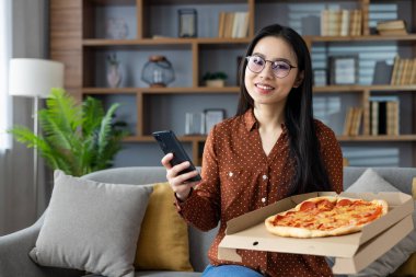 Asyalı kadın elinde telefonla koltukta oturmuş pepperonili pizza kutusuyla gülümsüyor. Rahat, rahat atmosfer rahatlık ve keyif verir..