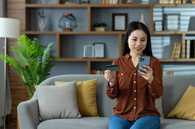 Evde elinde akıllı telefon ve kredi kartı olan Asyalı bir kadın online alışveriş yaparken gülümsüyor. Konforlu ortam, modern yaşam tarzı ve dijital işlemlerin kolaylığı vurgulandı.
