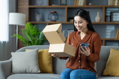 Kanepede oturan Asyalı kadın elinde akıllı telefonuyla paketi mutlulukla teslim ediyor. İnternetten alışveriş yapmanın ve evde sipariş almanın heyecanı. Neşeli boks deneyimini temsil eder.