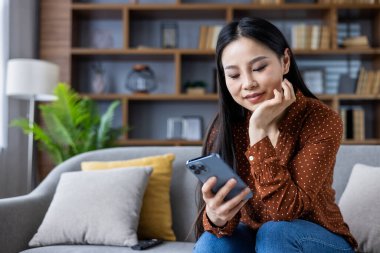 Sıradan giyimli Asyalı bir kadın koltukta oturmuş akıllı telefon kullanıyor ve rahatça gülümsüyor. Dekoratif unsurlarla modern ev dekorasyonu, kapalı alanlarda eğlenmek..