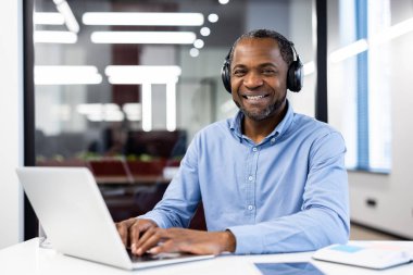 Ofiste dizüstü bilgisayar kullanırken gülümseyen yetişkin bir Afro-Amerikan işadamı. Kulaklık takmak modern teknolojiyi ve bağlantıyı akla getiriyor. Parlak ayar verimliliği ve rahatlığı iletir.