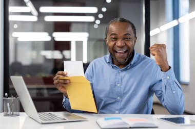 Mature African American businessman celebrates success at office desk with laptop and open envelope, displaying joy and accomplishment. Image conveys excitement, achievement, and career progress. clipart