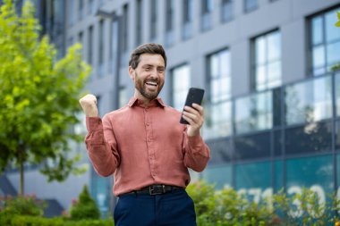 Neşeli, olgun iş adamı dışarıda akıllı telefon kullanarak kutlama yapıyor. Profesyonel kıyafetli bir adam neşe, başarı ve mutluluğu ifade ediyor. Teknoloji kullanımı ile vurgulanan olumlu duygular, iyimserlik yayıyor