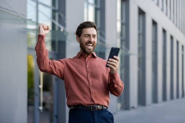 İş adamı akıllı telefona bakarken sevinçten havalara uçuyor. Mutlu olgun bir adam zaferle yumruğunu ofisin dışında kaldırıyor. Başarı kavramı, başarı, teknoloji, telefon iletişimi.